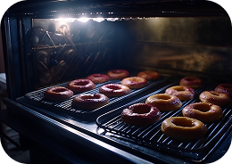 Donuts baking in a dimly lit oven, arranged on metal racks. The warm glow from the oven light highlights their golden texture, creating an inviting ambiance.