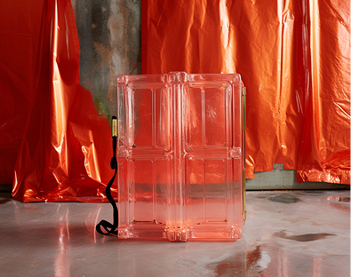 A transparent pink suitcase with a black strap stands on a polished floor. The background features shiny orange foil drapes, creating a modern, vibrant ambiance