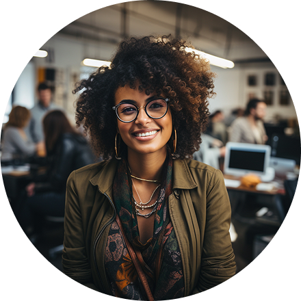 Smiling woman with curly hair and glasses in a casual outfit, sits in a modern, busy office space