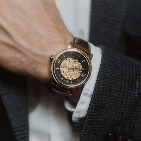 A close-up of a person wearing an elegant wristwatch with a gold skeleton dial and brown leather strap. The watch is paired with a navy suit, showcasing sophistication.